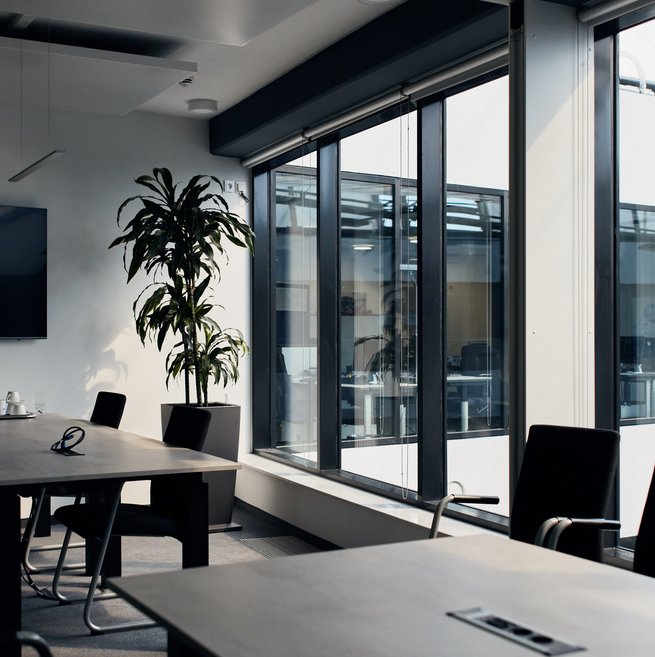 Moderner Büroinnenraum mit großen Fenstern, mehreren Tischen, schwarzen Stühlen und einer hohen Zimmerpflanze.