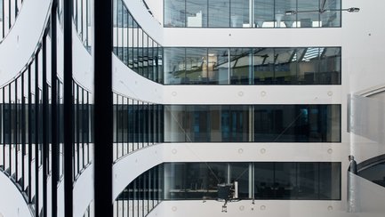 Innenraum eines modernen Bürogebäudes mit weißen Wänden, großen Fenstern und mehreren Etagen mit Glasgeländern.