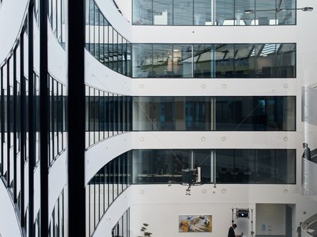 Innenraum eines modernen Bürogebäudes mit weißen Wänden, großen Fenstern und mehreren Etagen mit Glasgeländern.