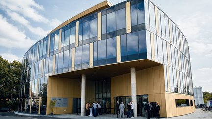 Modernes Bürogebäude mit großer Glasfassade und goldfarbenen Paneelen, Menschen stehen am Eingang.